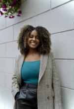 An individual stands in front of a cement block wall under purple flowers hanging above her head, smiling, wearing black pants, a blue shirt, and a plaid jacket