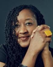 A headshot photo of LaShawnDa smiling with her hands clasped together in front of a dark gray background