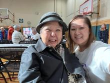 Two women sit next to each other in a gymnasium, one wearing a gray hat and button down shirt, and the other wearing a white t-shirt