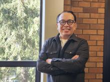 Dept. Chair Rick Bonus stands in front of a window and brick wall arms crossed wearing glasses and a black button-down shirt