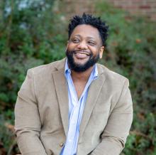 Oliver looks on smiling wearing a beige jacket over a blue button-down shirt