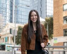 A woman stands smiling wearing a brown jacket and a purse slung over her left shoulder