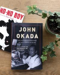 View from above of two books, NO-NO BOY and JOHN OKADA, next to a potted plant on a wooden tabletop