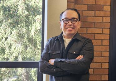 Dept. Chair Rick Bonus stands in front of a window and brick wall arms crossed wearing glasses and a black button-down shirt