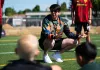 Han Eckelberg wearing a multi-color long-sleeve shirt, crouching in front of a group of children speaking to them