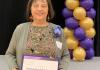 A woman stands in front of a stage holding a white parchment paper that reads Anjélica Hernández-Cordero 2024 distinguished staff award nominee. Behind her is a stack of purple and gold balloons.