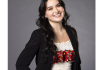 María stands in front of a gray background, smiling, wearing a white shirt with a floral pattern across the middle and a black blazer over the shirt