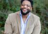 Oliver looks on smiling wearing a beige jacket over a blue button-down shirt