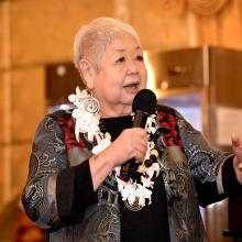 An Asian woman wearing a gray, black, red, and gold shirt with a lei over her neck, speaks to the audience with a microphone in her right hand