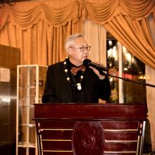 An Asian man wearing a black suit jacket, red and gold bow tie, with a necklace with an assortment of beads, speaks to the audience using a microphone attached to a mic stand